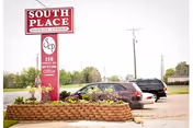 Outdoor view of the South Place Rehabilitation & Skilled Nursing facility sign with a landscaped flower bed in front. Several parked cars and utility poles are visible in the background under a clear sky.