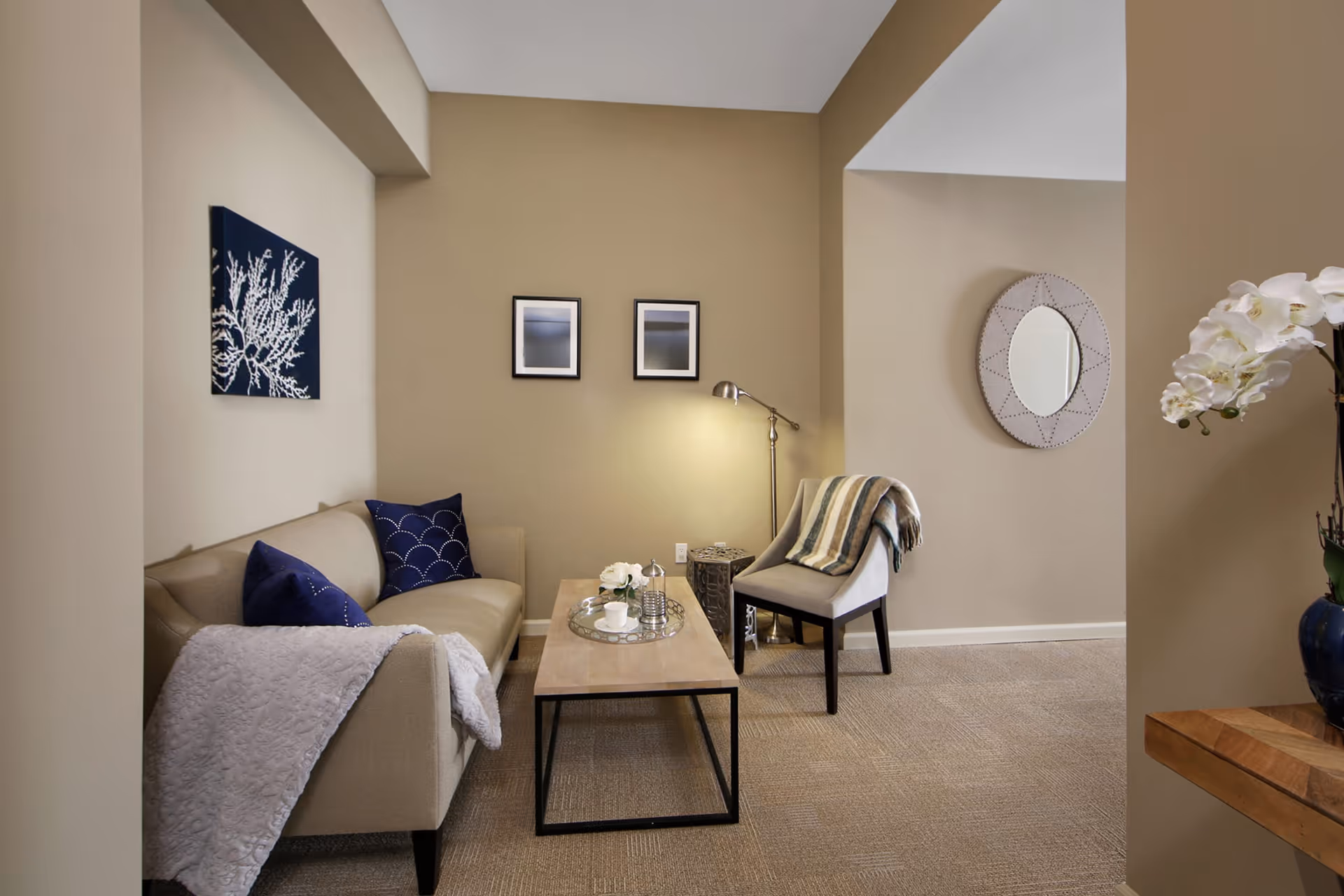 A cozy living room area with a beige sofa adorned with blue pillows and a white throw blanket. In front of the sofa is a wooden coffee table with a tray holding a teapot, cups, and a small vase with flowers. To the right, there is a beige upholstered chair with a striped throw blanket draped over it, next to a floor lamp. The walls are painted beige and decorated with two framed pictures and a round mirror. A vase with white flowers is partially visible on the right side.