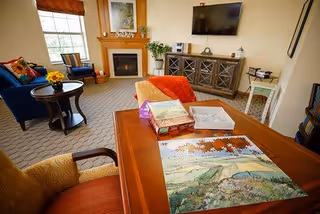 A cozy living room with a wooden table in the foreground featuring a partially completed jigsaw puzzle and puzzle boxes. The room includes a fireplace with a decorative 'believe' sign above it, a flat-screen TV mounted on the wall, a cabinet below the TV, and comfortable seating including an orange armchair and a blue armchair near a window.