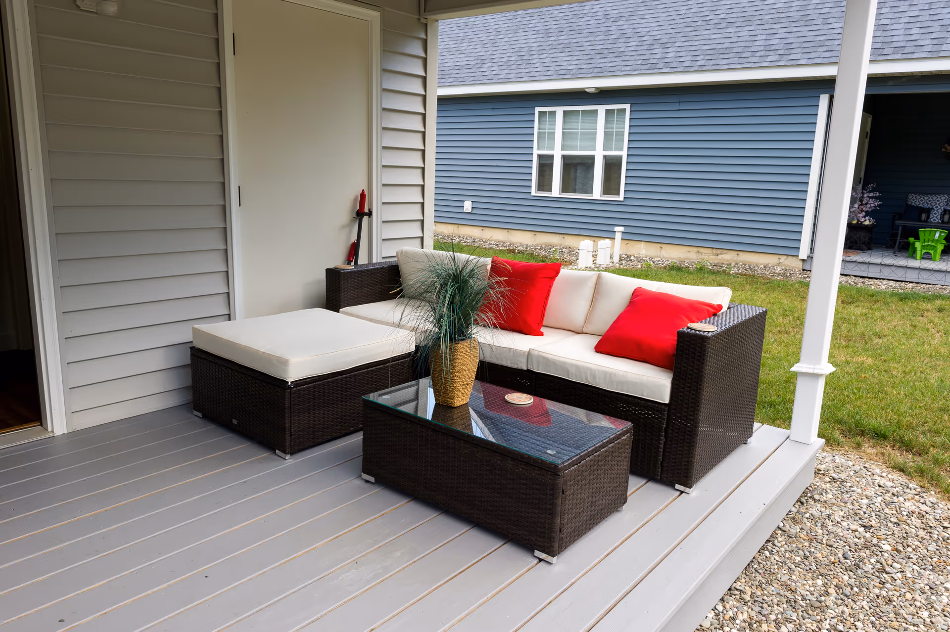 Wicker patio sofa with cream cushions and red pillows on a covered porch next to a glass-top coffee table and potted plant.