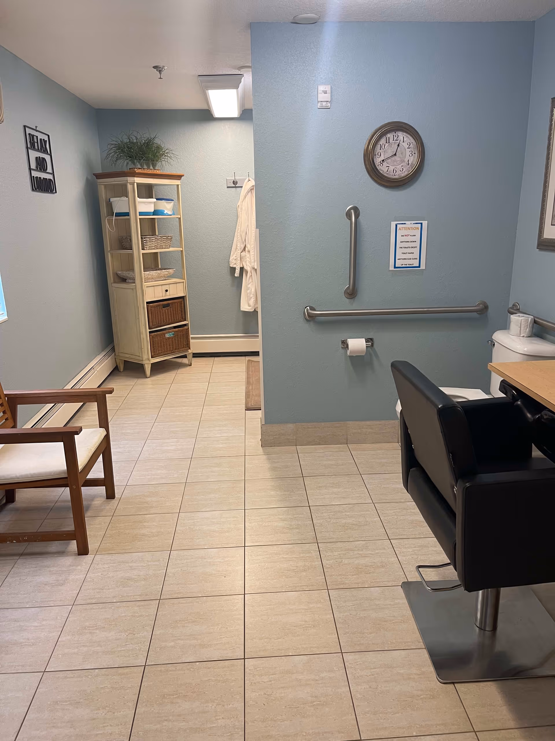 Interior room with light blue walls and beige tiled floor. On the right side, there is a black salon-style chair in front of a counter with a toilet next to it. A clock and a sign are mounted on the wall above grab bars and a toilet paper holder. On the left side, there is a wooden chair with a white cushion and a tall shelving unit with baskets and towels. A white robe hangs on a hook in the back corner. A framed sign on the wall reads 'RELAX AND UNWIND'.