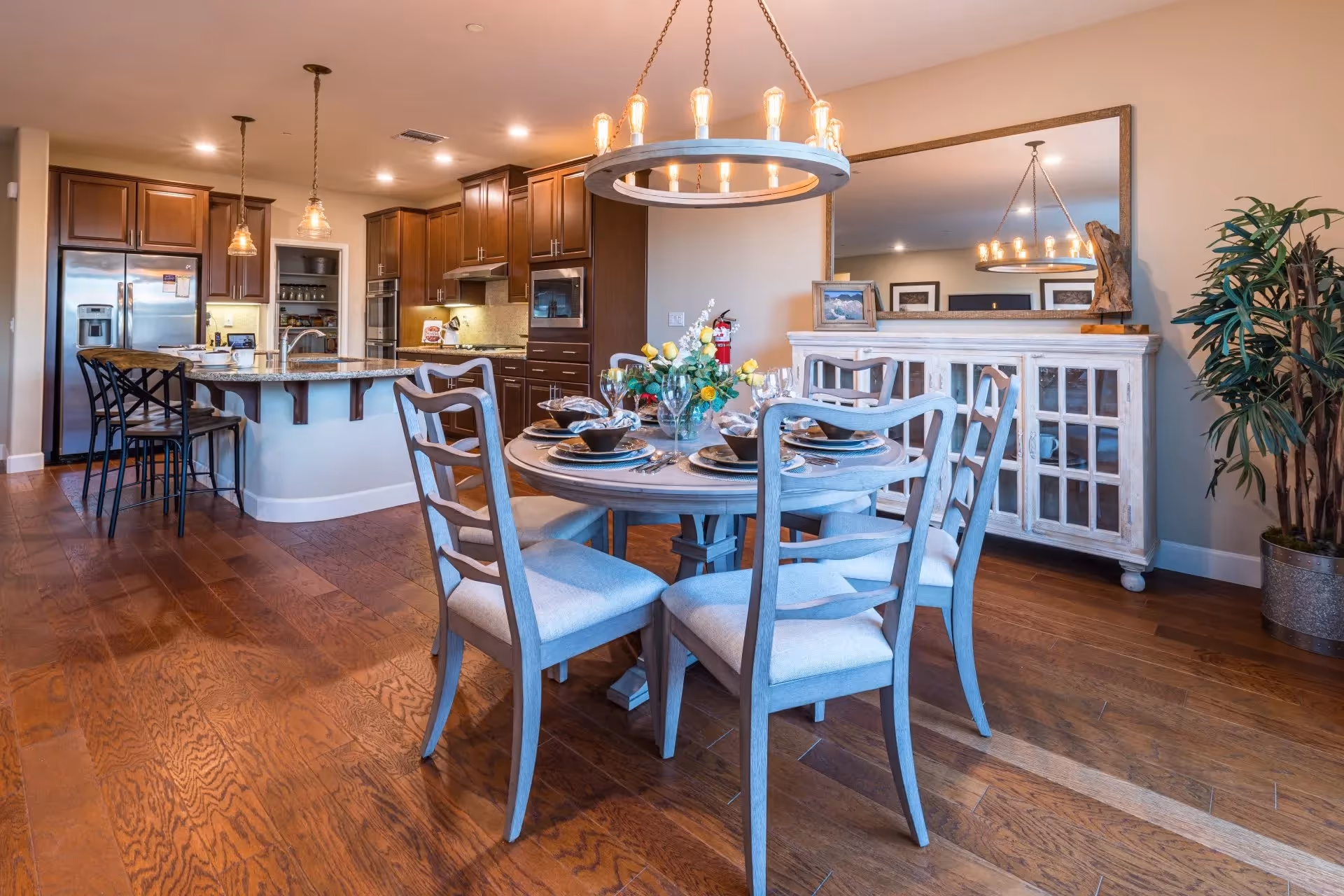 A bright and spacious dining area connected to a kitchen. The dining area features a round wooden table set with plates, bowls, glasses, and a vase of yellow and white flowers. Four cushioned chairs surround the table. The kitchen has dark wooden cabinets, a large island with a granite countertop, and three bar stools. Stainless steel appliances, including a refrigerator and oven, are visible. A large mirror hangs on the wall above a white sideboard, and a potted plant is placed in the corner. Warm wooden flooring extends throughout the space.