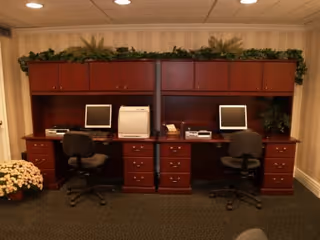 A room with two adjacent wooden desks, each equipped with a computer monitor, keyboard, and office chair. The desks have multiple drawers and overhead cabinets with green plants on top. There are additional office equipment items like a printer and a scanner on the desks. The room has beige wallpaper and a carpeted floor.