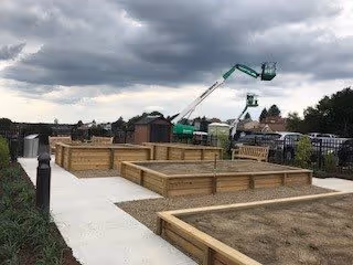 Outdoor garden area with raised wooden planter boxes along a concrete pathway under a cloudy sky. In the background, there is a green construction lift and some parked vehicles near a fence.