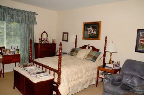A cozy bedroom with a wooden four-poster bed covered with a beige quilt and multiple pillows. There is a wooden chest at the foot of the bed with magazines and books on top. To the left, a window with green curtains and a small table with framed photos. A wooden dresser with a mirror and framed pictures is against the wall. On the right side, a wooden nightstand with a lamp and a gray upholstered armchair. The walls are decorated with framed artwork.