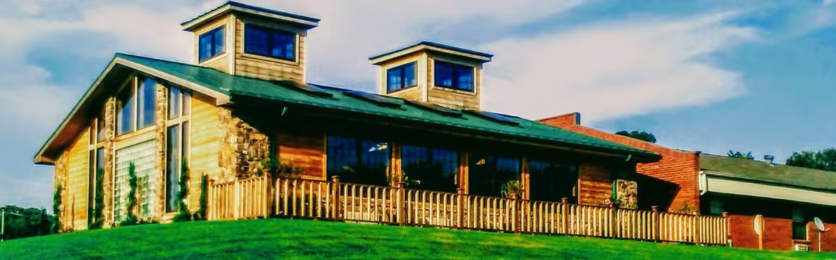 Exterior view of a building with a green roof, wooden siding, and large windows. The building has two small tower-like structures on the roof and a wooden fence along the front. The sky is partly cloudy and the building is situated on a grassy hill.