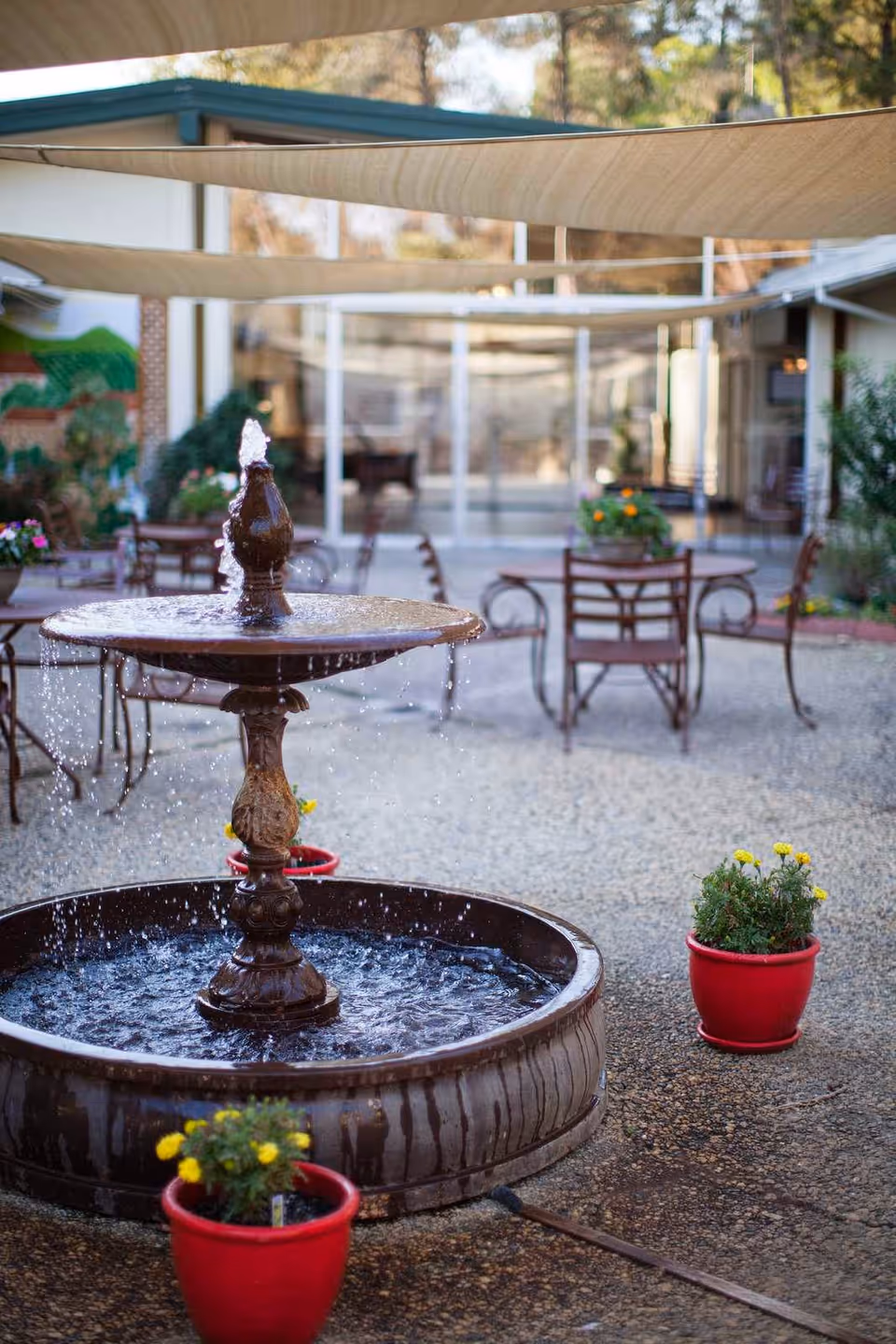 Outdoor courtyard area with a decorative water fountain in the center surrounded by red flower pots with yellow flowers. There are metal chairs and tables in the background under beige shade sails, with a building and glass doors visible behind.