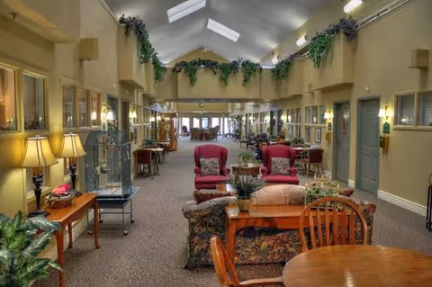 A spacious and well-lit common area in an assisted living facility featuring comfortable seating including armchairs and sofas, wooden tables and chairs, decorative plants, lamps, and a high ceiling with skylights. The walls are painted beige with several doors and windows along the sides.