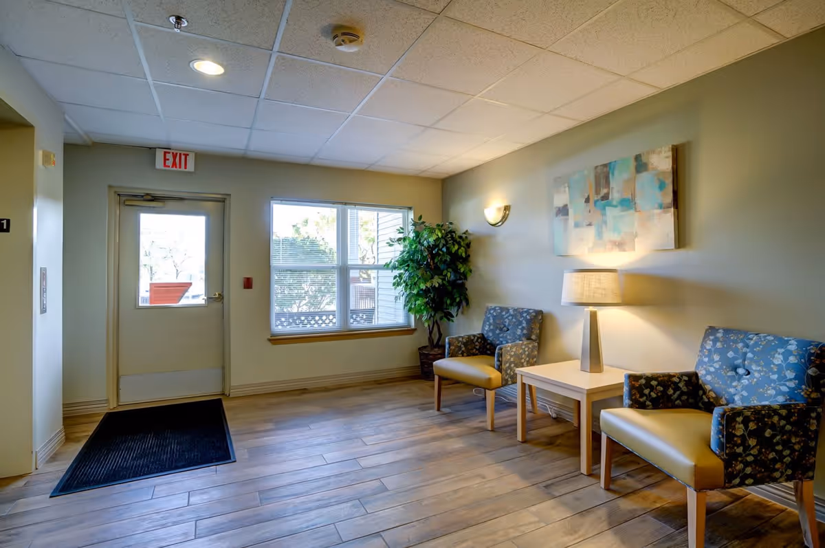 A small seating area inside a facility with two patterned armchairs, a wooden side table with a lamp, a potted plant, a window, and an exit door with an exit sign above it. The floor is wooden and the walls are painted light green with a piece of abstract artwork hanging above the chairs.