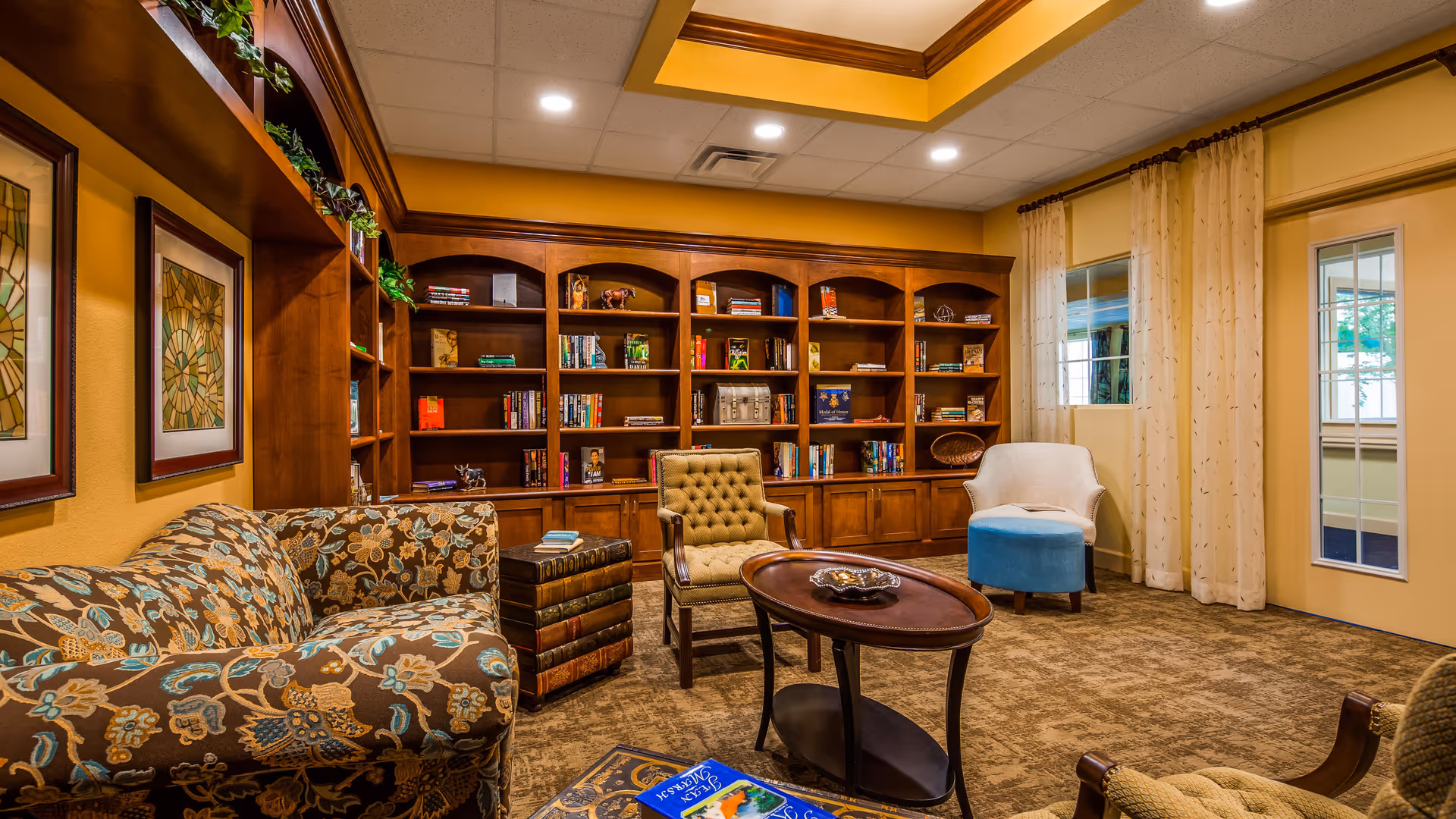 A cozy library or reading room with wooden bookshelves filled with books and decorative items, a floral patterned sofa, a tufted armchair, a white chair with a blue ottoman, a wooden coffee table, and framed artwork on the wall. The room has warm yellow walls and carpeted flooring.