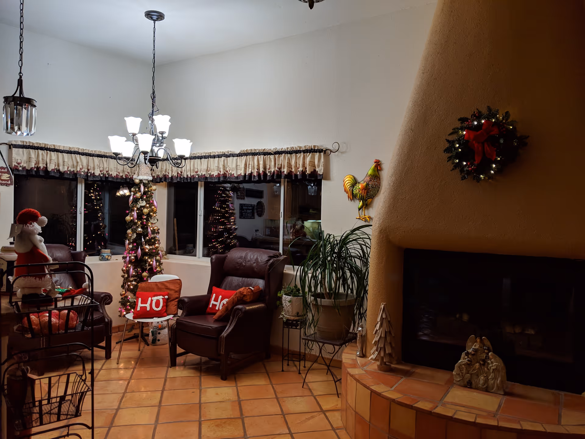 Cozy living room decorated for Christmas with a Christmas tree, two chairs with red pillows that say 'HO', a fireplace with a Christmas wreath above it, and various holiday decorations including a Santa figure and a rooster wall ornament.