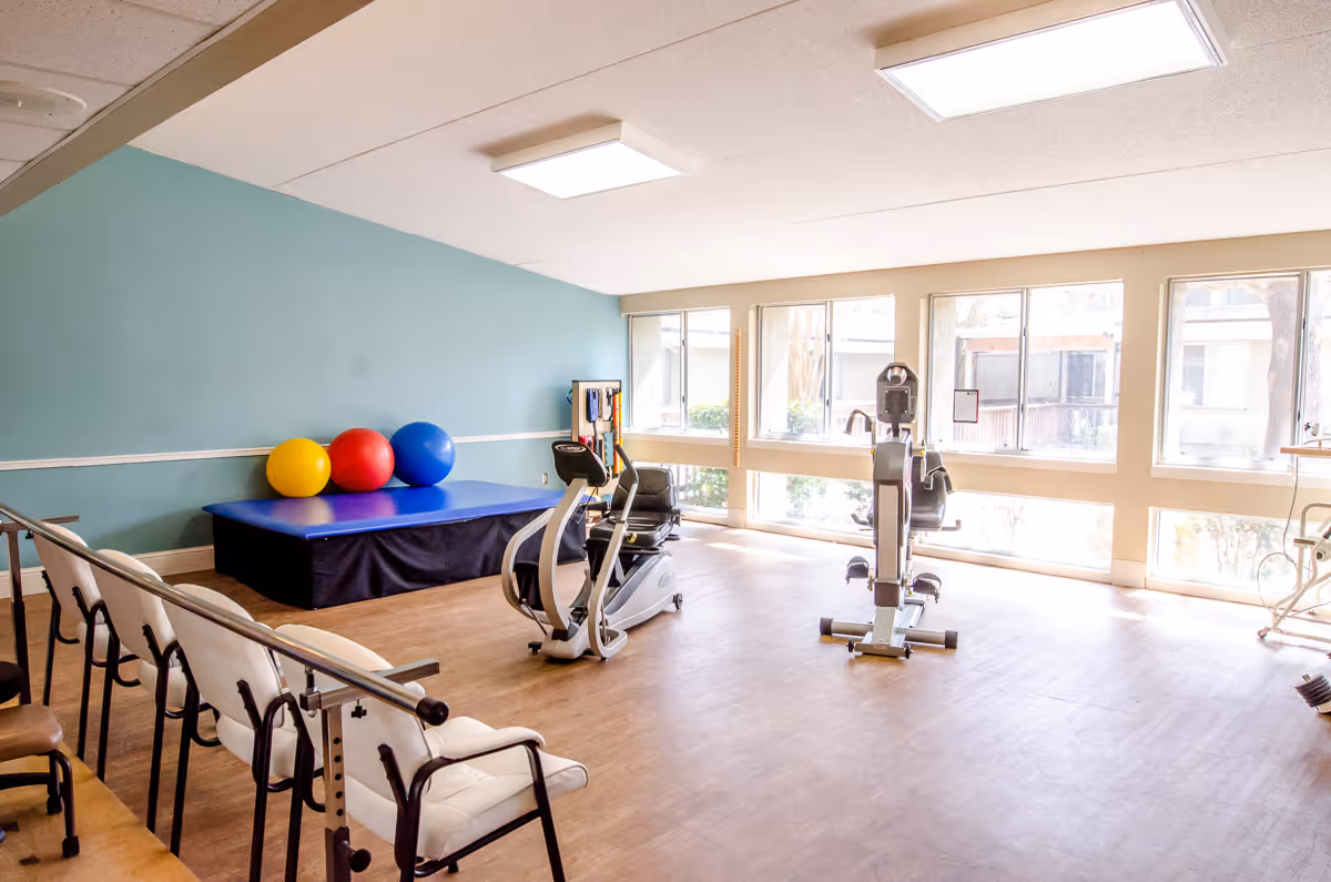 A bright exercise room with large windows letting in natural light. The room has a blue padded platform with three large exercise balls in yellow, red, and blue on top. There are two exercise bikes positioned near the windows and several chairs lined up along one side of the room. The walls are painted light blue and beige, and the floor is wooden.