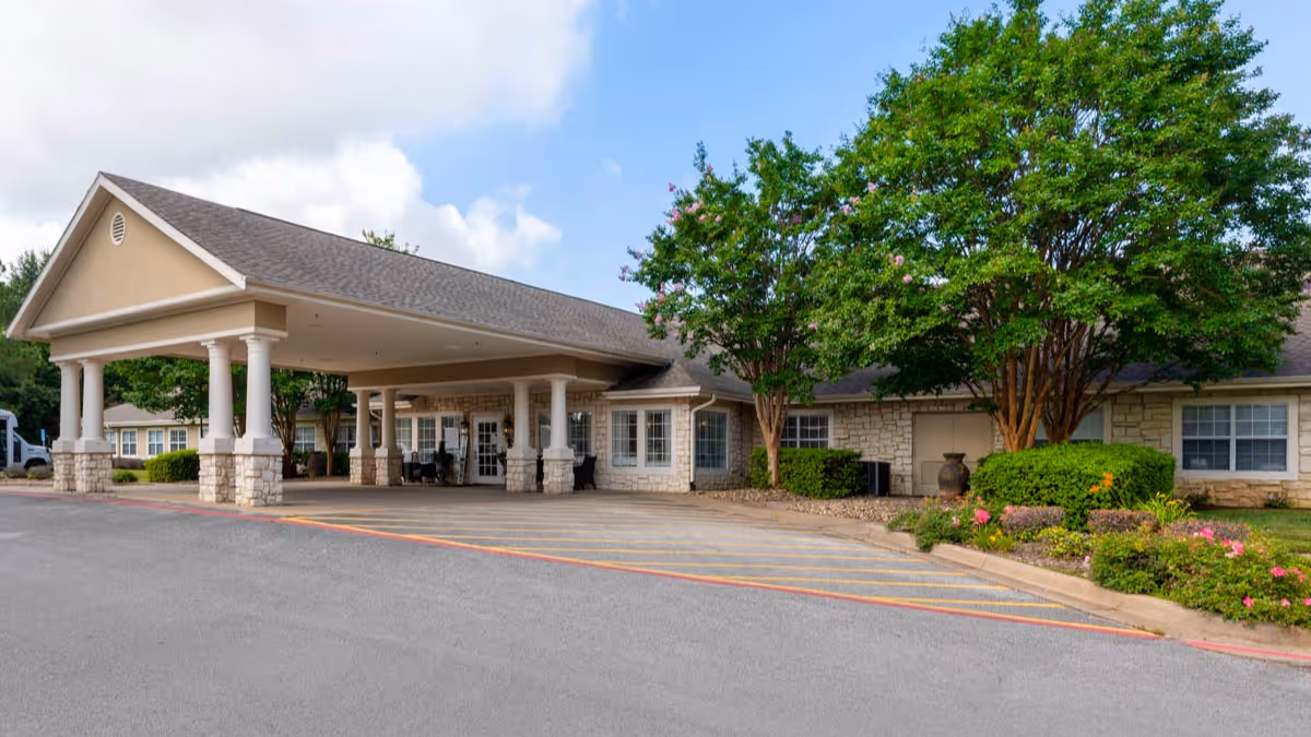 Exterior view of Brookdale Tyler South facility showing the main entrance with a covered drop-off area supported by columns, surrounded by landscaped greenery and trees under a partly cloudy sky.
