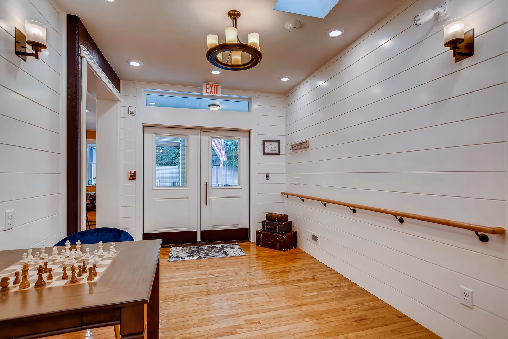 Interior hallway with white shiplap walls, wooden floor, and a wooden handrail on the right side. Double doors with windows lead outside, with an American flag visible through the glass. A small table with a chessboard and pieces is on the left side. Two stacked vintage suitcases are placed near the doors. Ceiling lights and wall sconces illuminate the space.