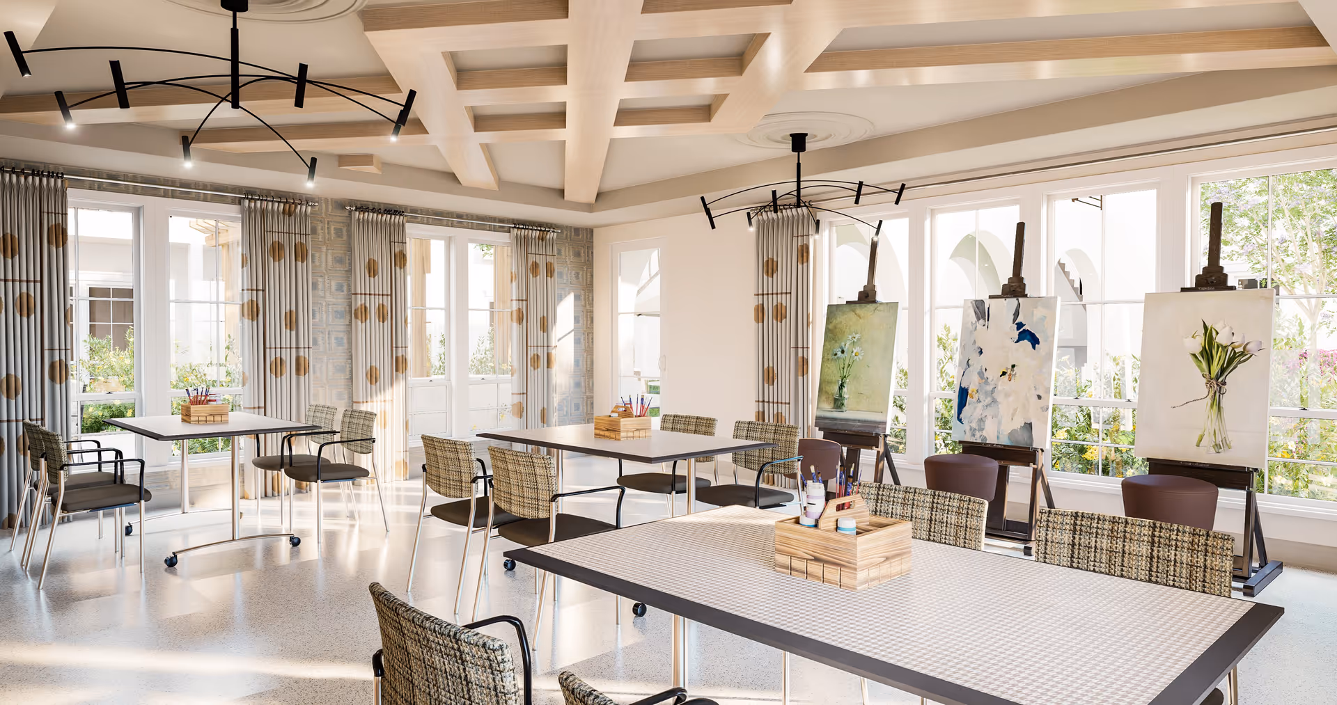A bright and spacious art room with large windows letting in natural light. The room features several tables and chairs arranged for group activities, with art supplies placed on the tables. Three easels display paintings of flowers near the windows. The ceiling has a decorative coffered design with modern light fixtures, and patterned curtains frame the windows.