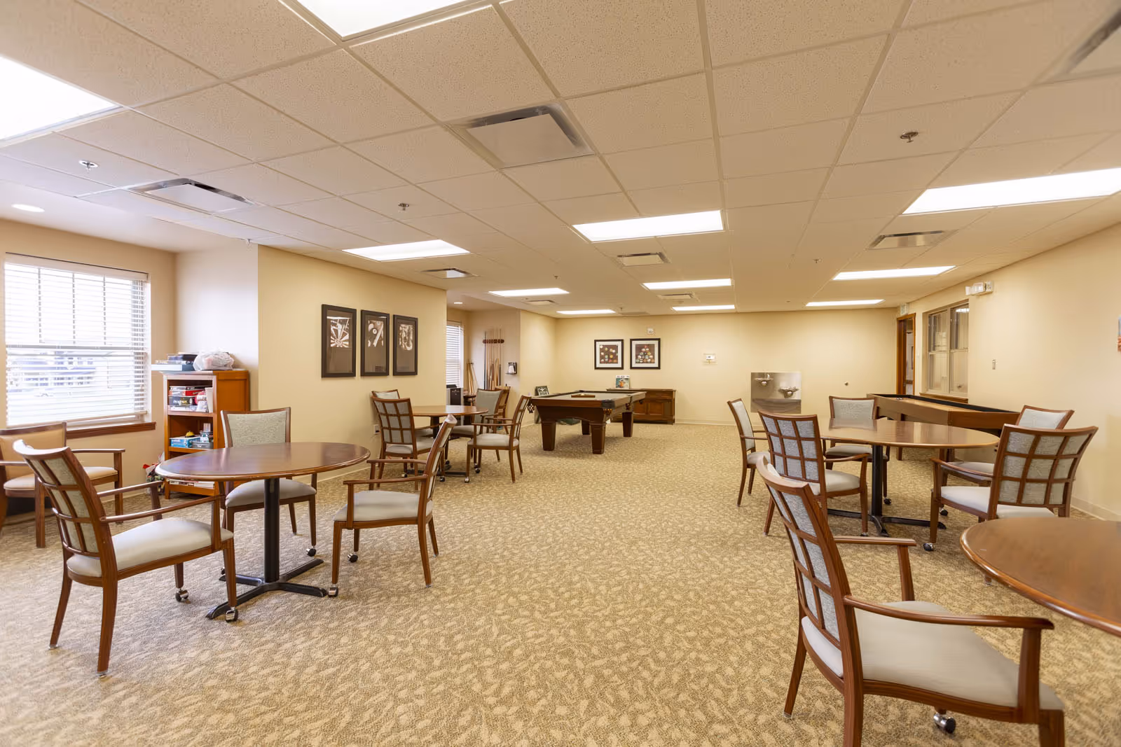 A spacious common area in a senior living facility with several round wooden tables and cushioned chairs arranged around the room. The room has beige walls, carpeted floors, and a ceiling with recessed lighting. There are framed pictures on the walls, a window with blinds, a bookshelf with games and books, and a pool table in the center back of the room.
