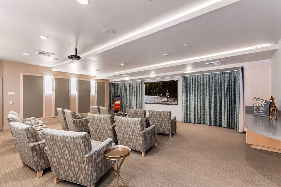 A cozy media room with two rows of patterned armchairs facing a large screen showing a nature scene. The room has beige walls with sound panels, recessed lighting, a popcorn machine in the corner, and green curtains covering the windows.