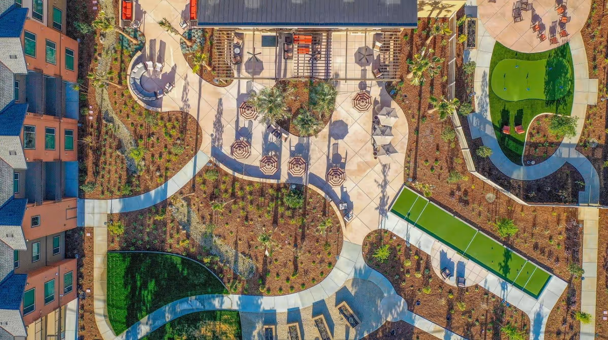 Aerial view of an outdoor recreational area at Bruceville Point featuring walking paths, landscaped gardens, seating areas with umbrellas, a small putting green, and a building with a shaded patio.