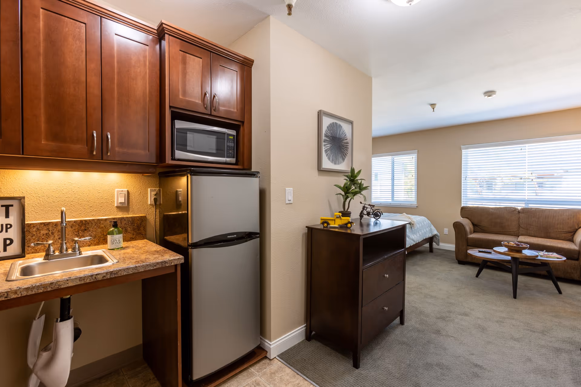 Interior view of a retirement community apartment showing a small kitchen area with wooden cabinets, a microwave, a mini refrigerator, and a sink. Adjacent to the kitchen is a living space with a brown sofa, a coffee table, a bed, and a dresser with decorative items. Large windows with blinds allow natural light into the room.