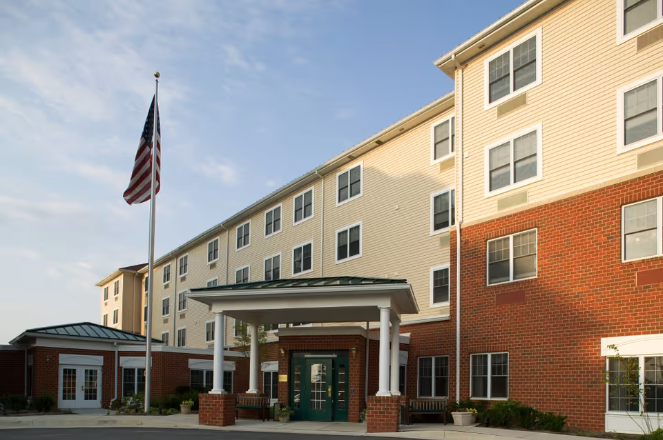 Exterior view of a multi-story senior living facility building with a brick and beige siding facade, a covered entrance supported by white columns, and an American flag on a flagpole in front under a partly cloudy sky.