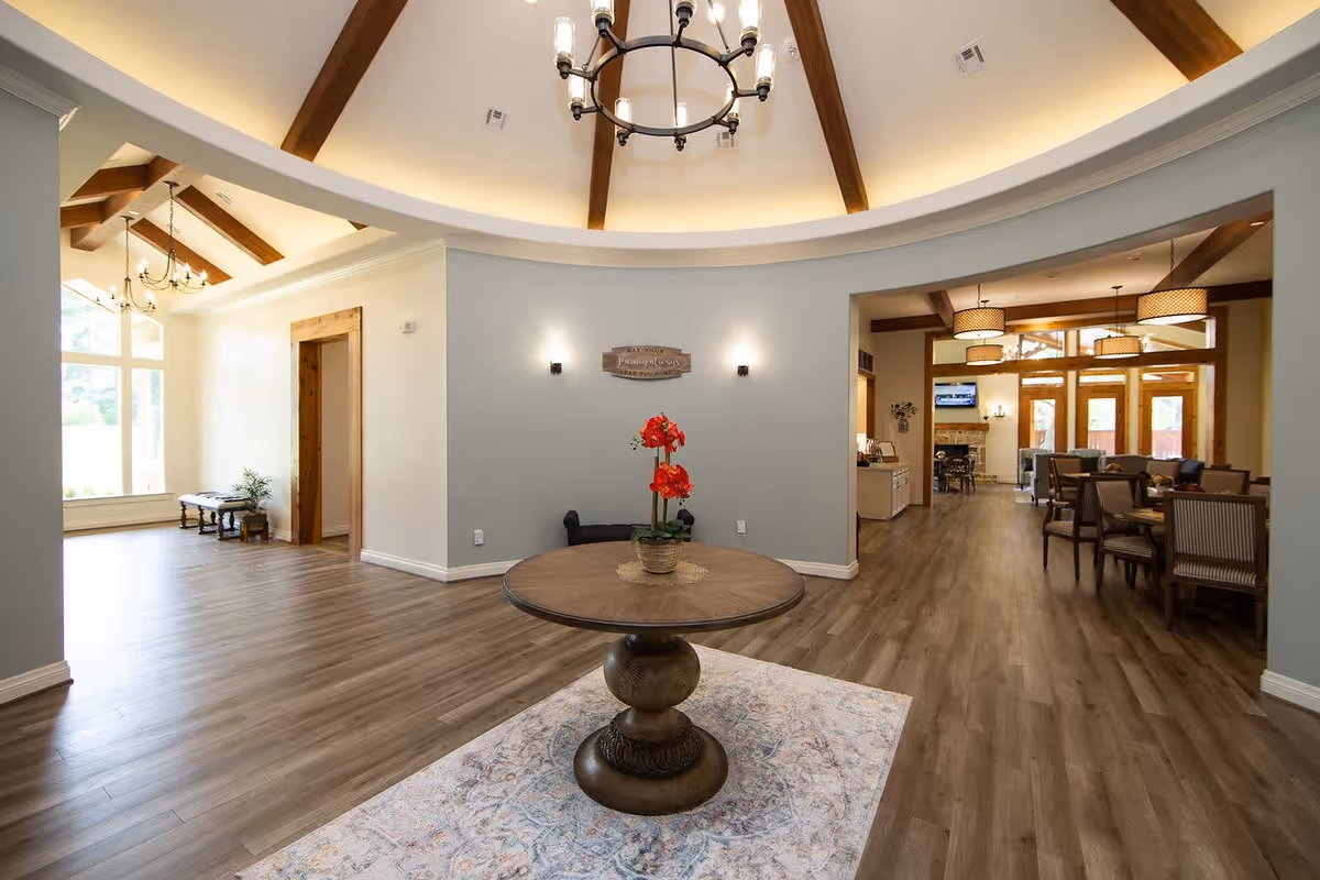 Interior view of a senior living facility with a round wooden table holding a red flower centerpiece on a patterned rug in the foreground. The room features wood flooring, a high ceiling with wooden beams and a chandelier, and soft lighting. To the right, there is a dining area with tables and chairs, large windows, and pendant lights. To the left, there is an open space with large windows letting in natural light.