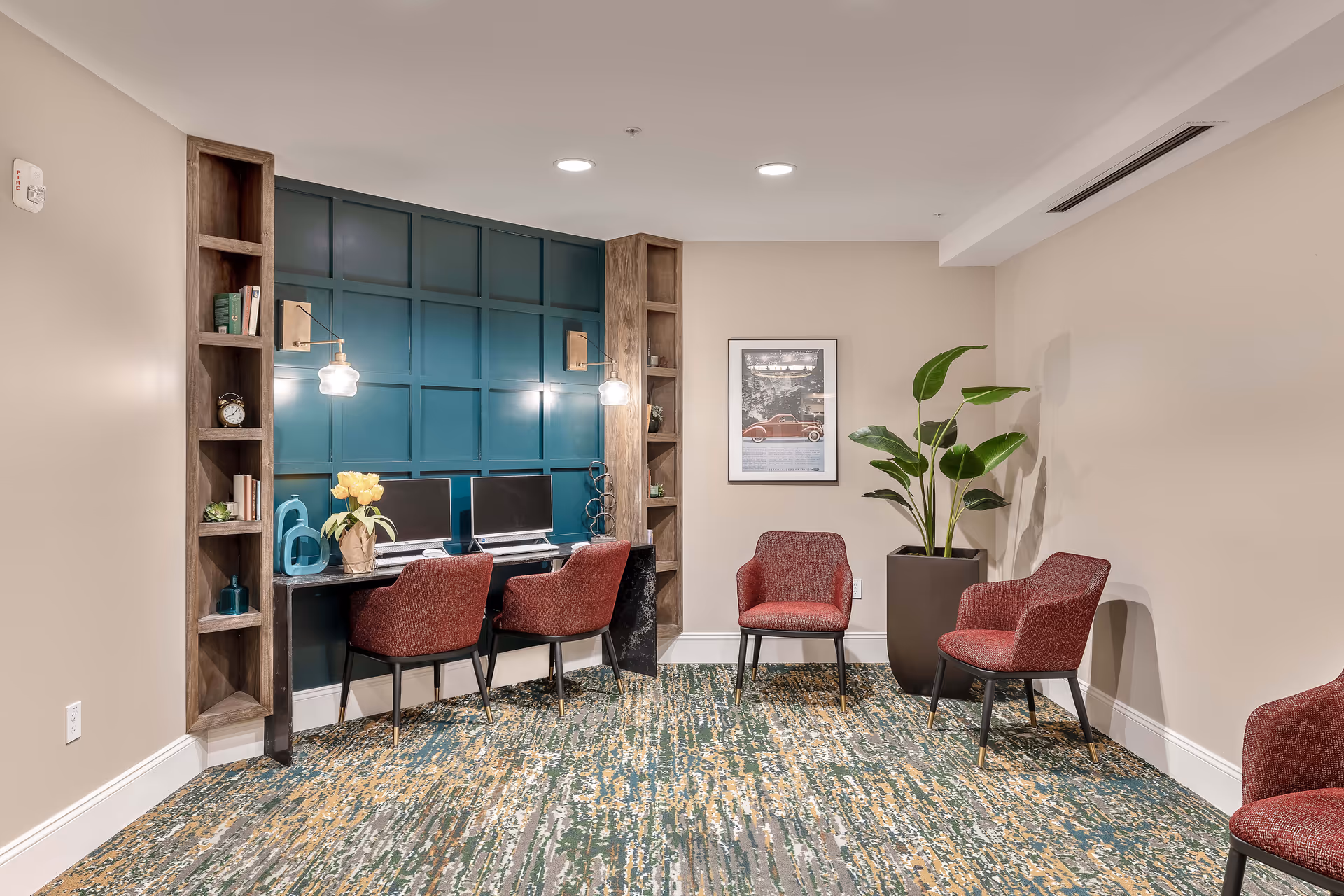 Communal seating area with a two-computer workstation set against a teal paneled wall, several red chairs, and a large potted plant.
