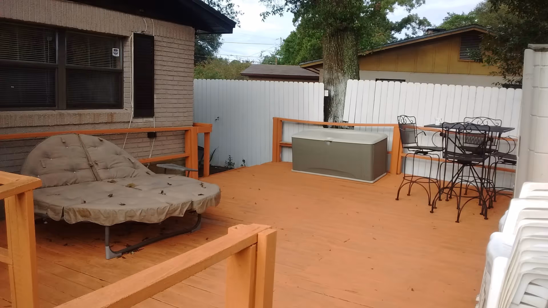 Wooden outdoor deck with a cushioned lounge chair, storage box, metal table and chairs beside a white fence and stacked plastic chairs.