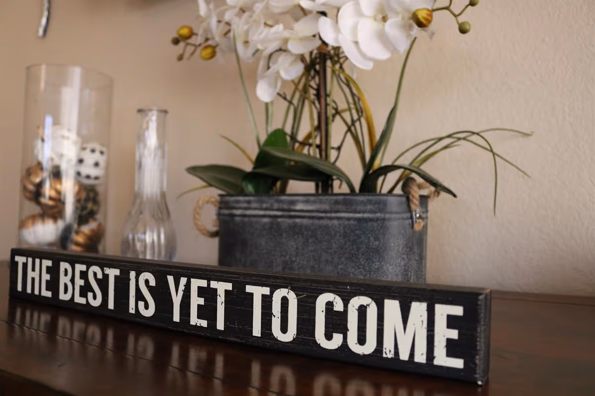 A decorative sign on a wooden surface with the text 'THE BEST IS YET TO COME' in white letters on a black background. Behind the sign, there is a metal container holding white orchid flowers and green leaves, and a glass vase filled with decorative balls.