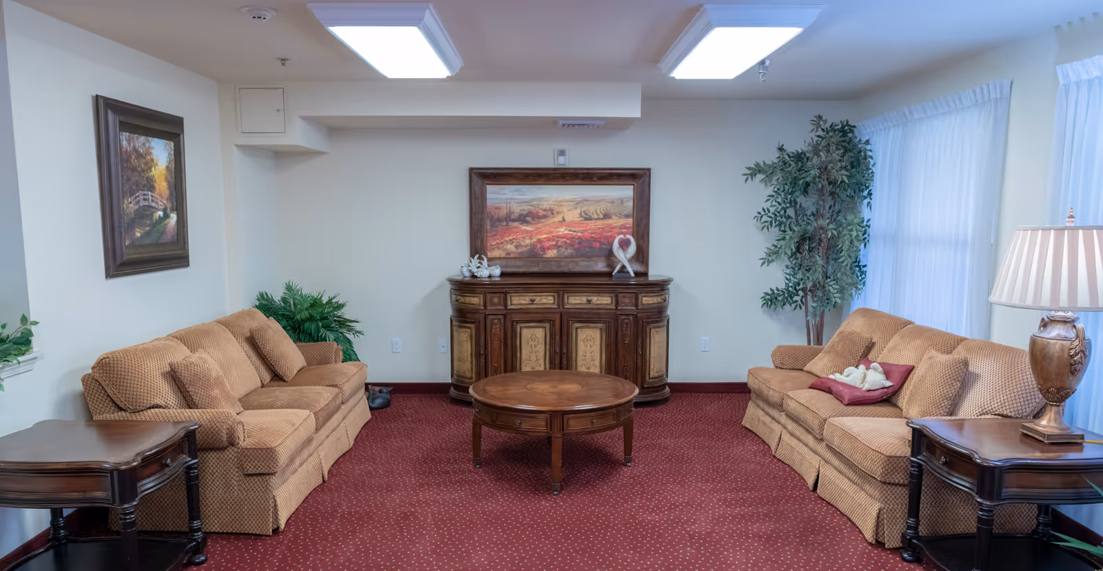 A cozy living room with two beige patterned sofas facing each other, a round wooden coffee table in the center, and a wooden sideboard against the back wall with a framed landscape painting above it. The room has red carpet flooring, white walls, potted plants, a table lamp on a side table, and large windows with sheer white curtains.