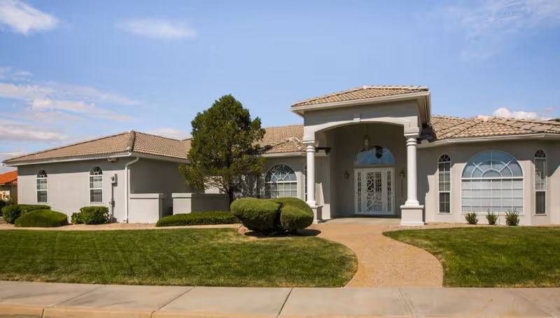 Front exterior view of a single-story assisted living facility building with a tiled roof, large arched windows, a covered entrance supported by columns, and neatly trimmed bushes and green lawn in front.