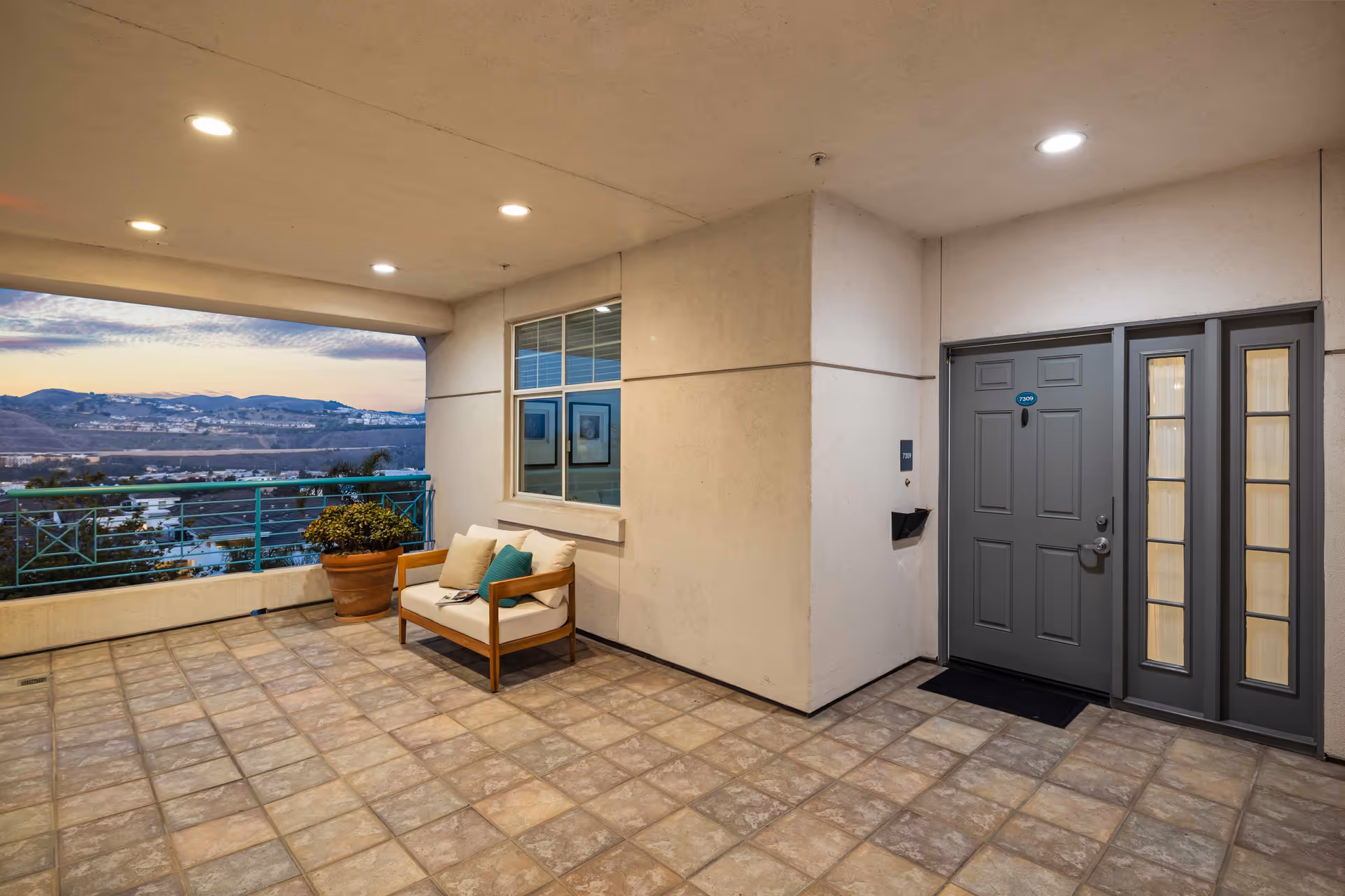 A spacious outdoor balcony area at the Sea Bluffs facility with tiled flooring, a cushioned wooden bench with pillows, a potted plant, and a gray entrance door with sidelights. The balcony has a railing and offers a scenic view of distant hills and a partly cloudy sky during sunset.