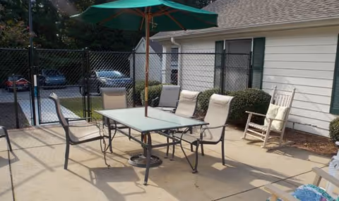 Outdoor patio area with a glass-top table surrounded by six chairs, including a rocking chair, and a green umbrella providing shade. The patio is adjacent to a white building with green shutters and bushes along the wall. A black chain-link fence separates the patio from a parking area with several parked cars.