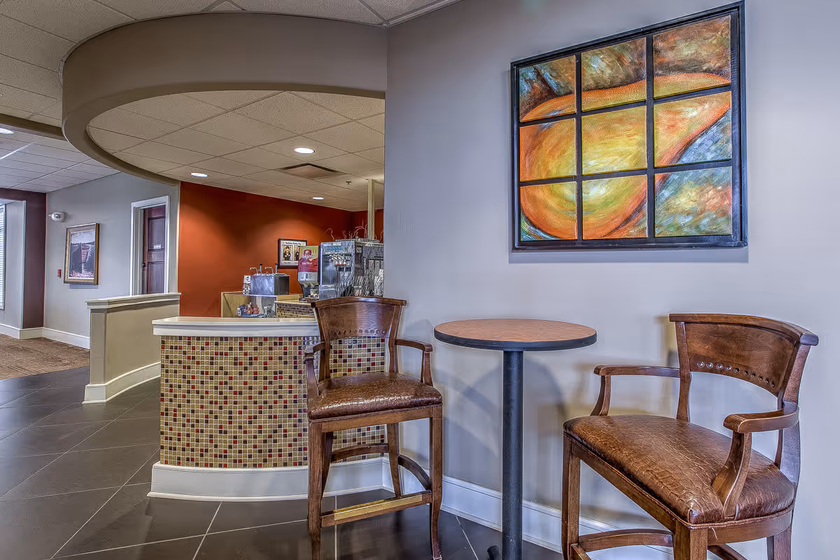 A seating area in an assisted living facility with two wooden chairs and a small round table between them. Behind the seating area is a curved counter with mosaic tile detailing and a coffee or beverage station. The walls are painted in neutral tones with a colorful abstract painting hanging above the chairs. The floor is tiled and the ceiling has recessed lighting.