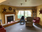 A cozy living room with a fireplace on the left wall decorated with stars and a wreath above it. In front of a large window with beige curtains, there is a wheelchair, a beige recliner chair, and a red armchair. A glass display cabinet is positioned in the corner to the right of the window.
