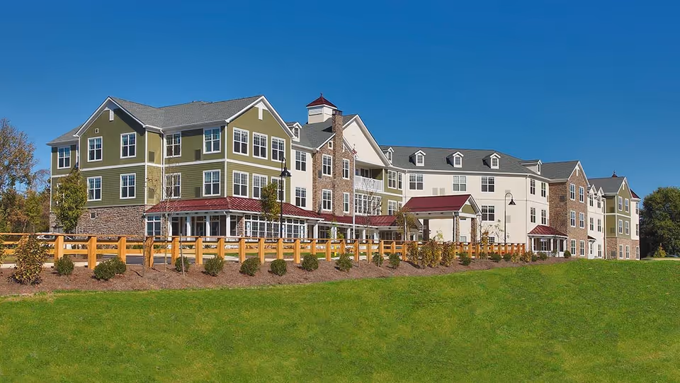 Exterior view of a large senior living facility building with green and beige siding, multiple windows, a stone facade, and red roofs. The building is surrounded by a wooden fence, landscaped bushes, and a green lawn under a clear blue sky.