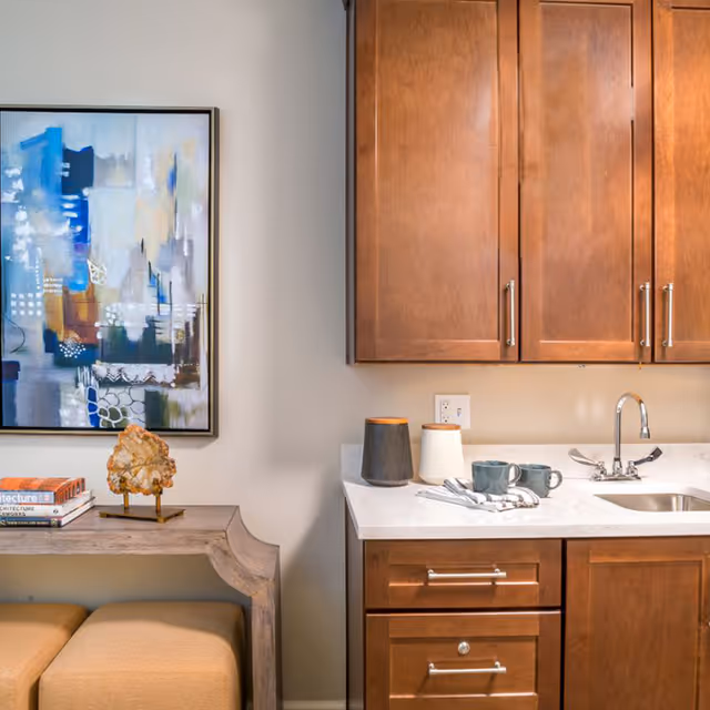 A small kitchen area with wooden cabinets, a white countertop with a sink and faucet, two mugs, and two containers. To the left, there is a wooden console table with two books, a decorative rock, and two beige ottomans underneath. A colorful abstract painting hangs on the wall above the console table.