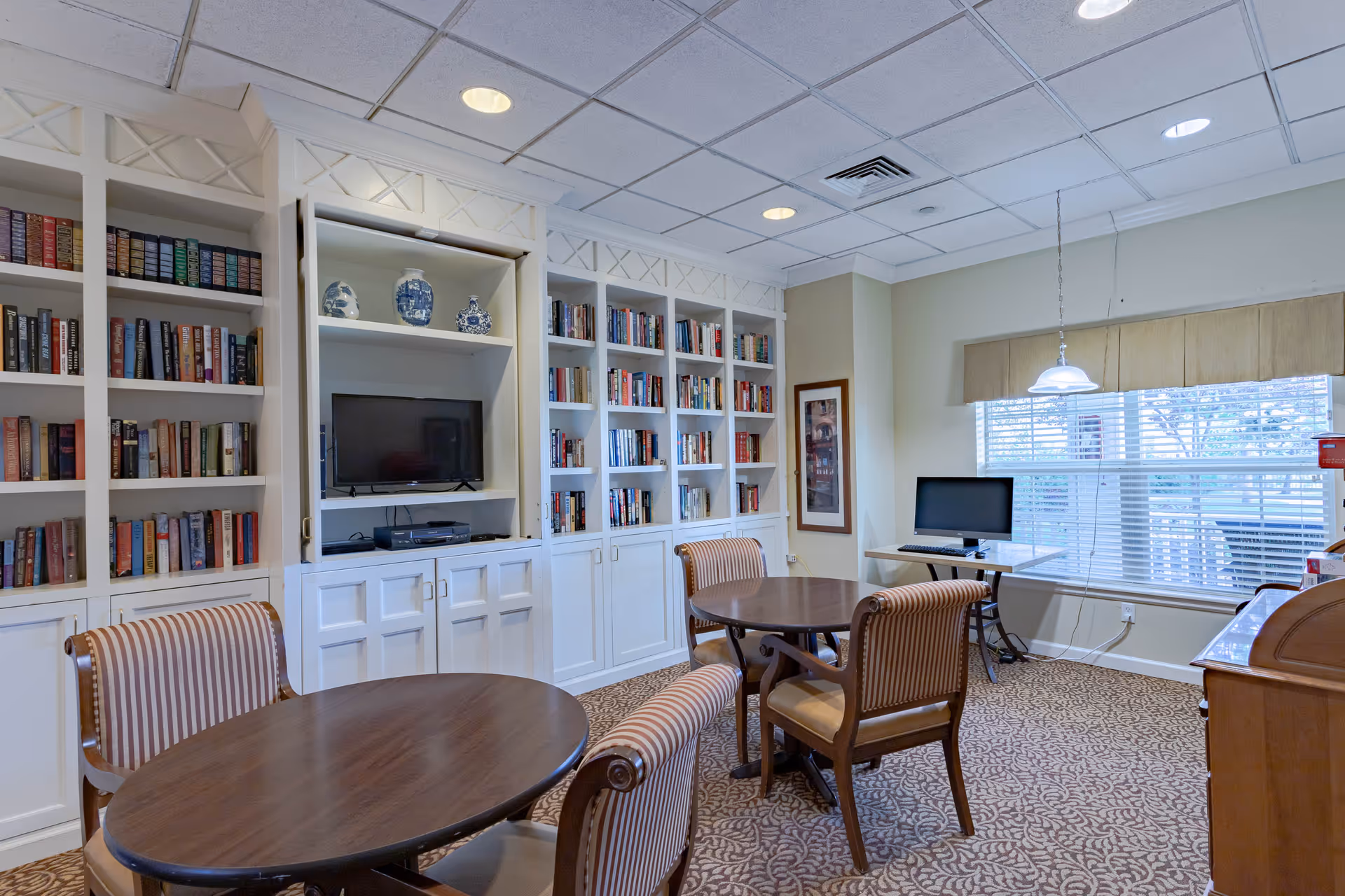 A cozy common room with built-in white bookshelves filled with books and decorative blue and white vases. There is a flat-screen TV and media player in the center of the shelves. The room has two round wooden tables surrounded by striped upholstered chairs. A window with blinds lets in natural light, and a computer workstation is set up near the window. The ceiling has recessed lighting and a hanging light fixture above one table.