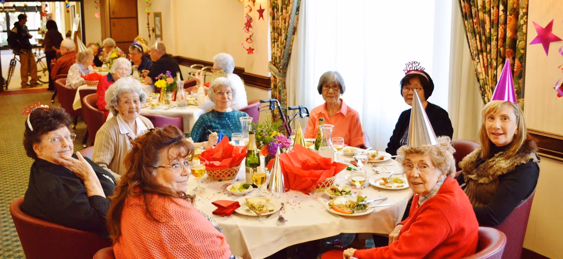 A group of elderly residents wearing party hats seated around a decorated dining table in a communal dining room.