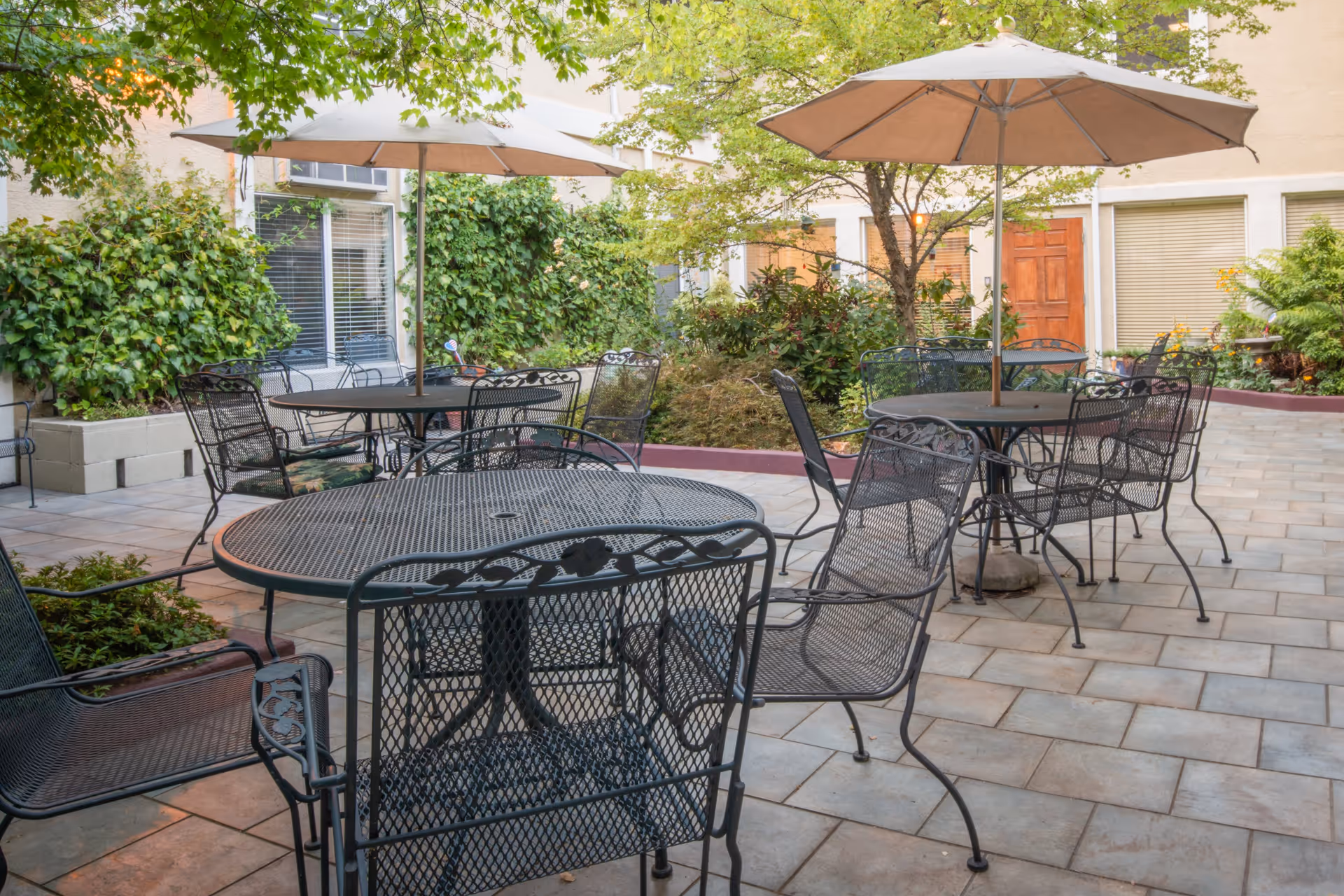 Outdoor patio area with multiple black metal tables and chairs, each table shaded by a large beige umbrella. The patio is surrounded by greenery including bushes and trees, with a building wall featuring windows and doors in the background.