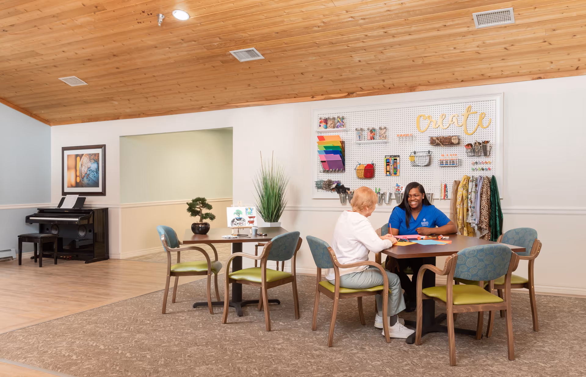 Two women sitting at a table in a well-lit activity room with a wooden ceiling. One woman is elderly and the other appears to be a caregiver or staff member. They are engaged in a creative activity with colorful paper and supplies. The wall behind them has a pegboard with various craft materials and the word 'create' displayed. There is a piano and framed artwork in the background.