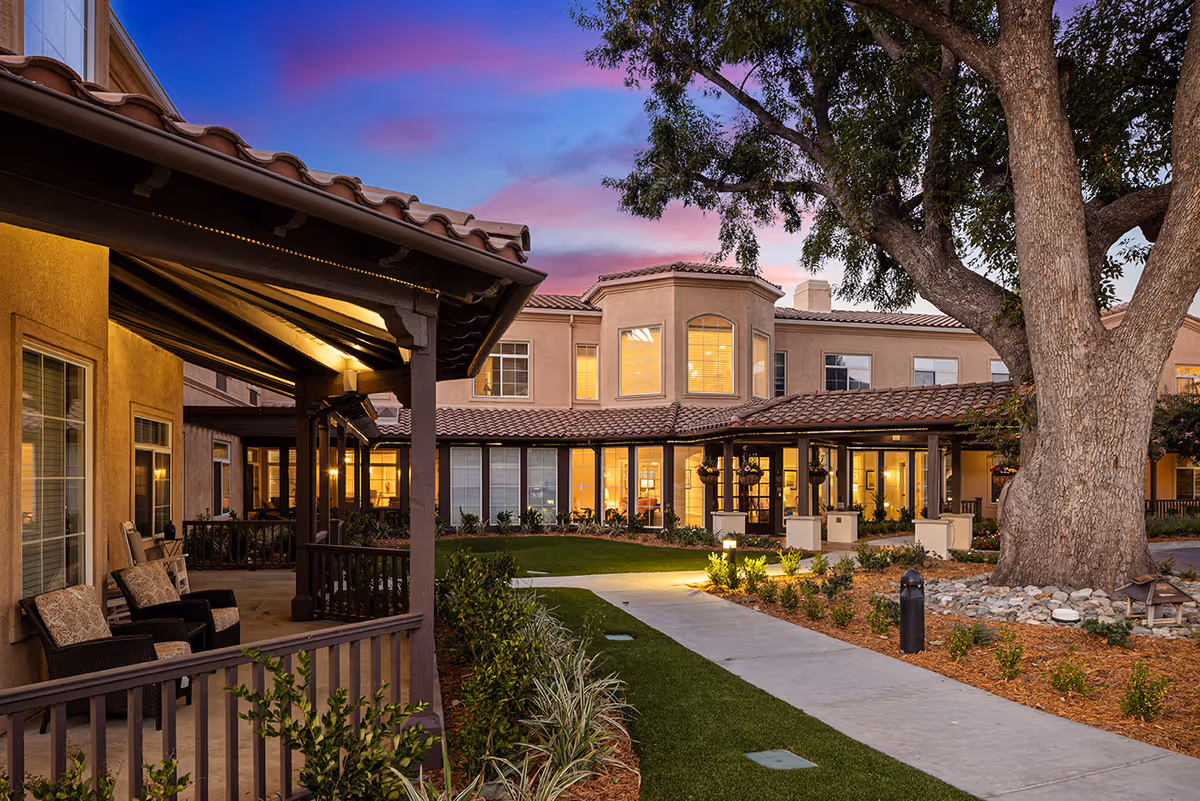 Well-lit courtyard and covered porch seating area in front of a two-story residential building at dusk.
