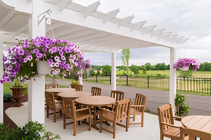 Outdoor patio area with wooden tables and chairs under a white pergola. Hanging baskets with vibrant purple flowers are suspended from the pergola. Green fields and trees are visible in the background under a cloudy sky.