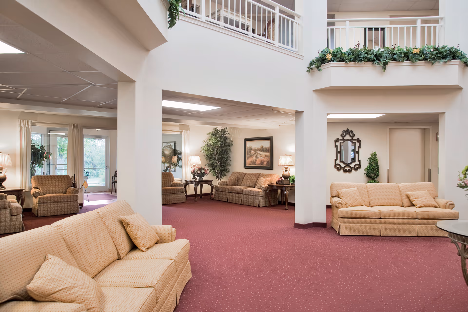 Open seating lounge with beige sofas and armchairs, side tables, lamps and plants beneath an indoor balcony.
