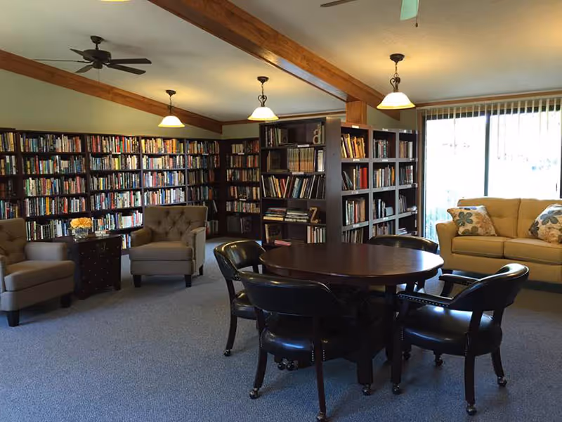 A cozy library room with multiple bookshelves filled with books along the walls. There are two beige armchairs with a small table between them, a round wooden table surrounded by four black chairs, and a beige sofa with floral pillows near a large window with vertical blinds. The ceiling has wooden beams and hanging light fixtures.