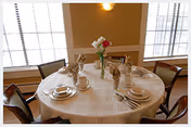A round dining table set for six people with white tablecloth, plates, cups, napkins folded in a decorative manner, and a vase with flowers in the center. The room has large windows with blinds and beige walls with white trim.