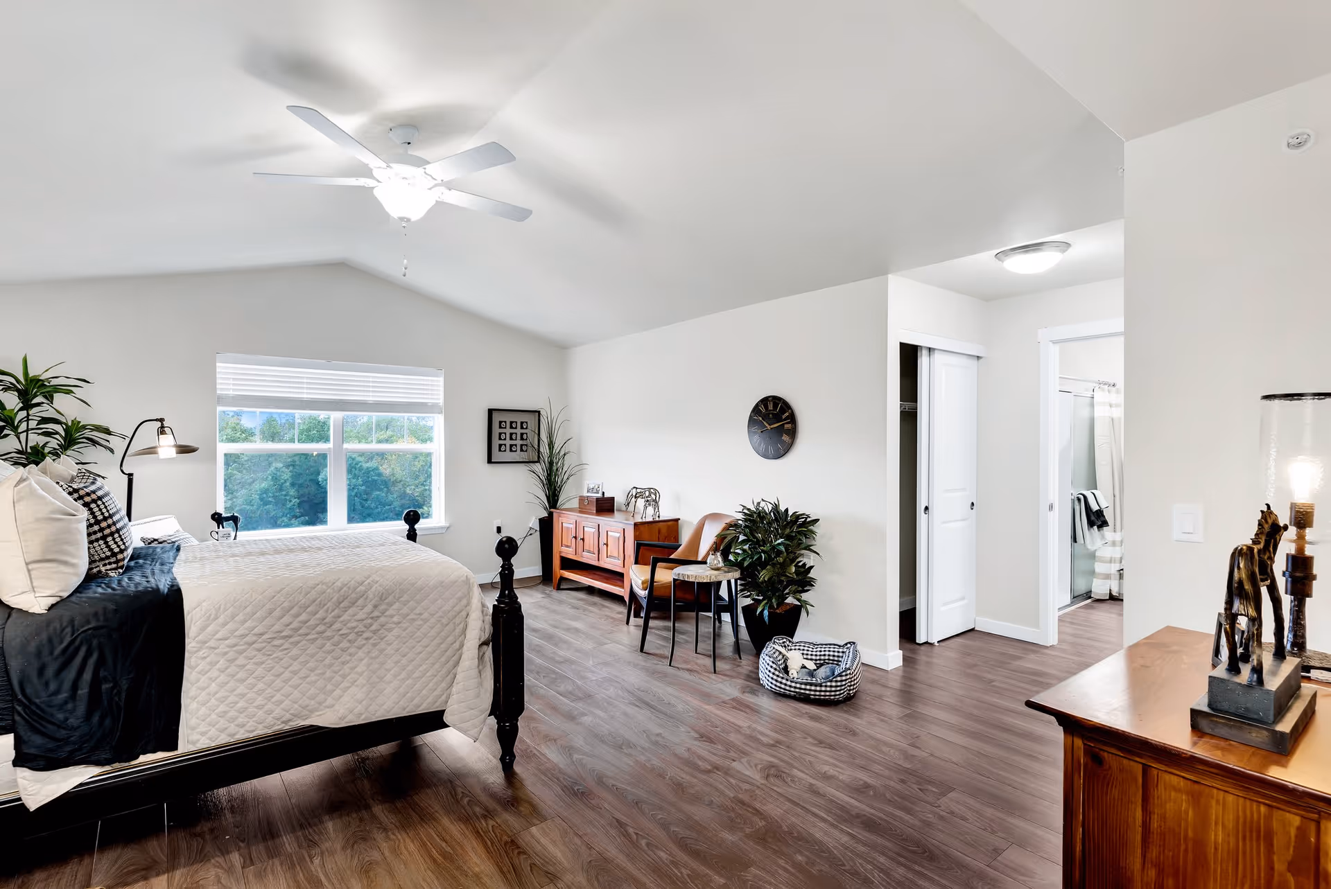 A bright and spacious bedroom with a large window showing greenery outside. The room features a bed with white and black bedding, a ceiling fan with light, wooden flooring, a wooden dresser with decorative items, a leather chair with a small side table, a potted plant, and an open doorway leading to a bathroom with a shower and towels.