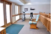 A small exercise area in a nursing home with two stationary exercise bikes, a wooden rocking chair, a green mat on the floor, large windows, and a wooden railing along one side.