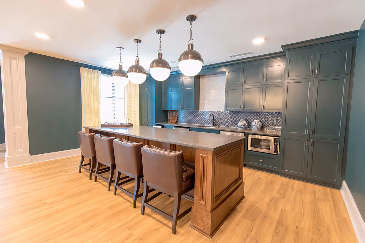 Modern kitchen with dark green cabinetry, a large island with a wooden base and gray countertop, four brown leather chairs, pendant lights hanging above the island, light wood flooring, and a window with yellow curtains.