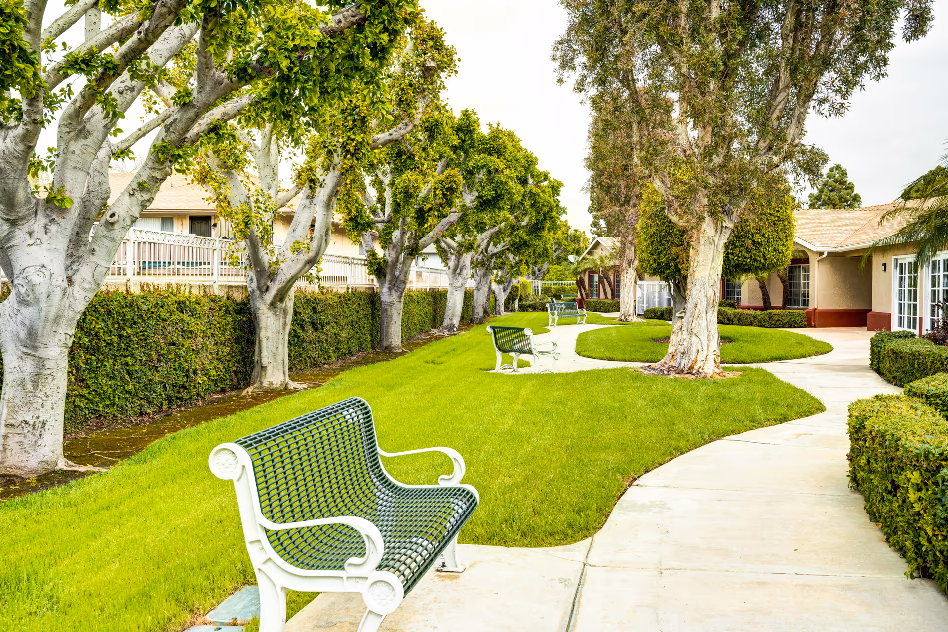 A landscaped outdoor area with a curved concrete walkway, green grass, several white and green metal benches, and trees lining the path. Residential-style buildings with beige walls and red trim are visible in the background.