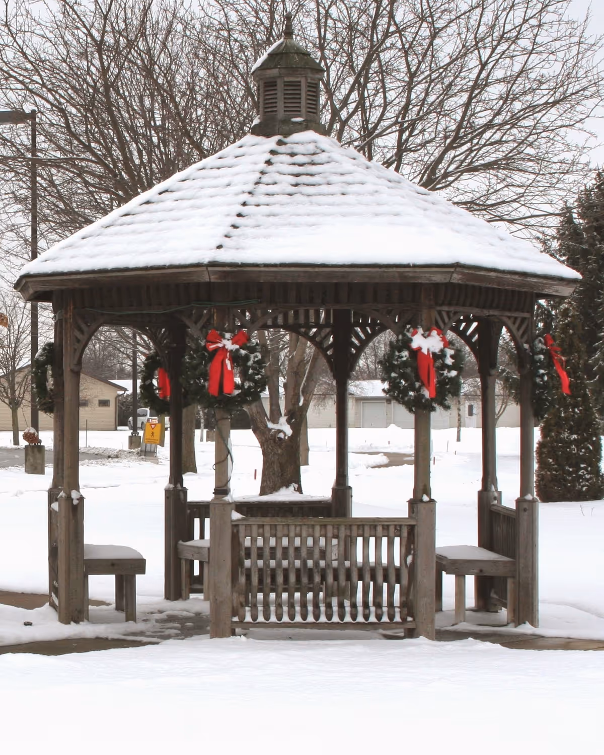 A wooden gazebo covered with snow on the roof, decorated with green wreaths adorned with red bows, surrounded by snow-covered ground and leafless trees in the background.